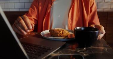 A stylish young man with glasses uses a laptop and eats a donut sitting at a table in a cafe, close-up of a man's hands, no face video
