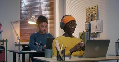 A man and a woman use laptops working at desks in the office. A man in a hat and headphones is talking on a chat video