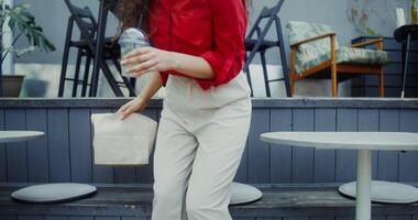 A young woman in casual clothes sits down at a table in a cafe, drinks a mojito from a disposable cup with a straw and takes a burger out of a paper bag video