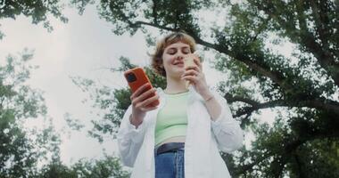 A young woman uses a mobile phone and eats ice cream while standing in the park on a sunny day video