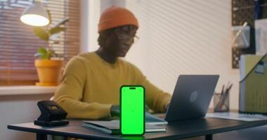 uma homem dentro informal roupas é digitando em uma computador portátil sentado às uma escrivaninha dentro a escritório. uma Móvel telefone com verde tela é em a mesa dentro frente do ele video