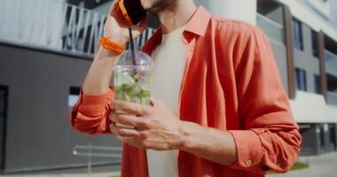 A young stylish man with glasses drinks mojito from a disposable cup with a straw and talks on a mobile phone while walking along a city street, no face video