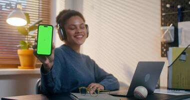 uma mulher dentro fones de ouvido sorrisos olhando às a Câmera e mostrando uma Móvel telefone com verde tela enquanto sentado às dela escrivaninha dentro a escritório video