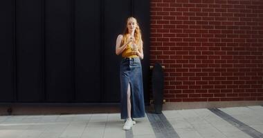 A young red-haired woman drinks lemonade from a disposable cup with a straw and smiles while looking at the camera, standing on a city street video