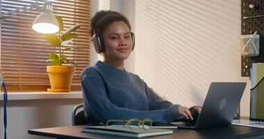 uma mulher vestindo fones de ouvido é sorridente às a Câmera sentado às uma escrivaninha dentro frente do a aberto computador portátil dentro a escritório video