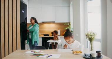 The girl enthusiastically uses a graphic tablet while sitting at the table. Mom gets the baby yogurt from the refrigerator in the background video
