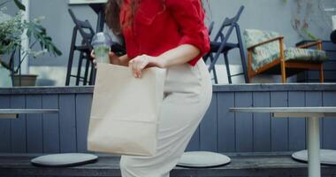 A young woman in casual clothes with a paper bag in her hand sits down at a table in a cafe, and drinks a mojito from a disposable cup with a straw video
