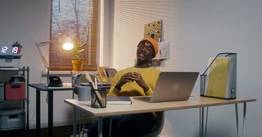 A man in a hat, dressed in casual clothes, smiles looking at the camera sitting in front of an open laptop in the office video