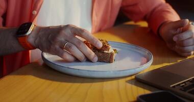 A stylishly dressed man eats bruschetta and uses a laptop while sitting at a table in a cafe, a close-up of men's hands, no face video