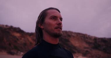 Close-up of the face of a young man in a diving suit, with wet hair, standing near a rock in the open air at dusk video