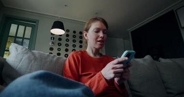 A young red-haired woman in casual clothes laughs while typing on a mobile phone while sitting on a sofa in a lotus pose. Home interior video