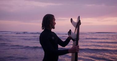 A man adjusts his diving suit standing on the seashore with a surfboard in his hand in the rays of the setting sun video