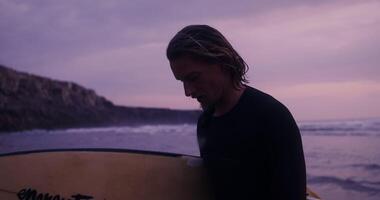 A man with a serious look walks along the seashore with a surfboard in his hands at sunset video