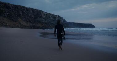 A man with a surfboard in his hands walks along the seashore, leaving footprints in the wet sand video