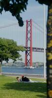A man and a woman are lying on a blanket on the grass, resting near the river. A red suspension bridge connects the banks of the river video