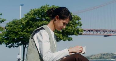 A young beautiful woman takes a book and starts reading sitting on a blanket near the city embankment with a red bridge over the river video