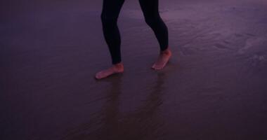 A man walks on wet sand on the seashore with a surfboard in his hands, after surfing at sunset video