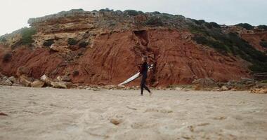 A young man in a diving suit pushes a surfboard into the sand on the beach near the red rocks video