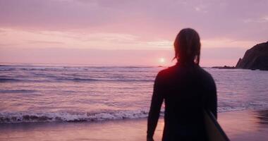 Rear view of a man in a diving suit with wet hair and a surfboard in his hands, looking into the sea at sunset video