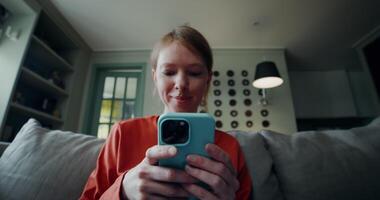 A young woman in casual clothes smiles typing on a mobile phone while sitting on a sofa in a home interior. The is focused on the phone video