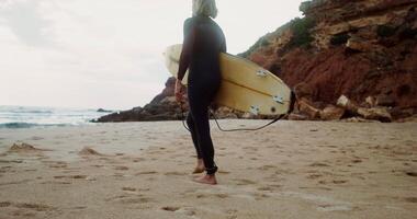 A man in a diving suit, with a surfboard in his hands, walks along a sandy beach towards the sea, rear view video