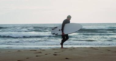 A man in a diving suit with a surfboard in his hand walks along the shore at dusk. Foam waves hit the sandy shore video