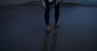 A man with a surfboard in his hands walks along the seashore at dusk, the moves up from the men's legs video