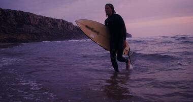 A young man in a diving suit, with a surfboard in his hands, comes out of the sea to the sandy shore, in the rays of the setting sun video