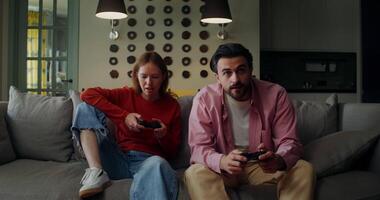 Stylishly dressed man and woman smile while playing games using joysticks while sitting on the couch at home video