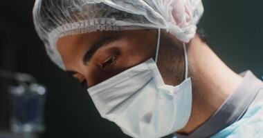 Close-up of a doctor's face in a medical mask and a disposable cap on his head, bent over a patient, focused while working in the operating room. in 4k, red komodo video