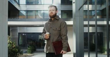 A bearded man in glasses, holding a disposable cup and a laptop in his hands, standing in the courtyard of office center and looking at camera with a smile. in 4k, red komodo video
