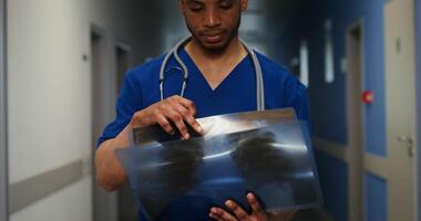 An African-American doctor in a medical uniform with a phonendoscope around his neck examines x-rays of the patient's lungs while standing in the corridor of the clinic. in 4k, red komodo video