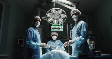 Three medics dressed in sterile medical clothing look at the camera while standing around an operating table with a patient lying on it in a modern operating room. in 4k, red komodo video