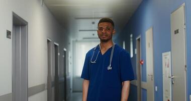 A doctor of African American appearance with a phonendoscope around his neck smiles while looking directly into camera while standing in a bright corridor of a medical center. in 4k, red komodo video