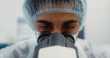 A male scientist in a disposable cap and a medical mask looks into the microscope. Close-up of the laboratory assistant's face, front view. 4k , red komodo video