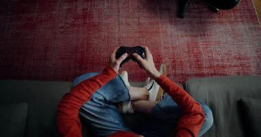 A woman in casual clothes is playing games with a joystick, sitting on a sofa in a lotus position, top view, no face video