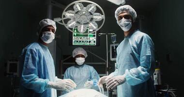 Three surgical medics look at the camera while standing around an operating table with a patient lying on it in a modern operating room. in 4k, red komodo video