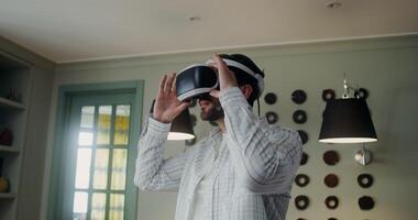 A stylish man in casual clothes puts on a virtual reality headset, and starts moving his hands standing in the middle of a room at home video