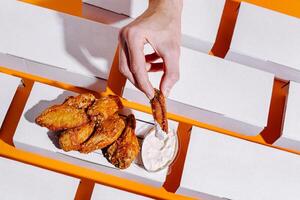 Hand dipping crispy chicken wings into creamy sauce on a bright orange surface with white boxes in the background photo