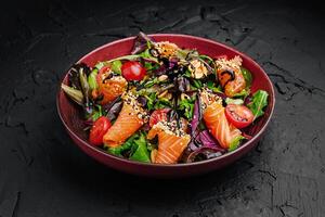 Fresh salmon salad featuring mixed greens, cherry tomatoes, and sesame seeds served in a red bowl on a dark textured background photo
