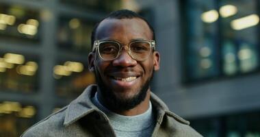 Stylish young African-American man smiling looking at the camera while standing near a modern building in the evening. Close-up of a man's face with glasses. in 4k, red komodo video