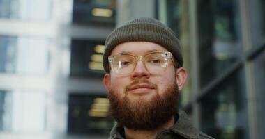 A young bearded man in a knitted tucked-up hat and glasses smiles while looking at the camera while standing outdoors near the building, close-up. in 4k, red komodo video