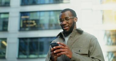 A stylish young African-American man smiles broadly while using a mobile phone, standing near a building in which lamps shine in the windows in the evening. in 4k, red komodo video