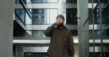 A young bearded man in a knitted tucked-up hat with a laptop in a case in his hands walks near an office building and drinks coffee from a disposable cup. in 4k, red komodo video