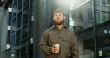 A young bearded stylishly dressed man looks at the camera, standing near an office building and holding a disposable cup, dynamic , the man's gaze moves behind the camera. in 4k, red komodo video