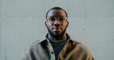 Young warmly dressed African-American man in transparent glasses smiling looking at the camera while standing against a gray concrete wall. in 4k, red komodo video