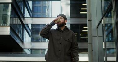 A young bearded man in a knitted hat drinks coffee from a disposable cup while walking near a stylish building with large windows, holding a laptop in a case in his hand. in 4k, red komodo video