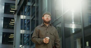 A young bearded man in a knitted hat looks at the camera, standing near a building with large stained glass windows and holding a disposable cup. in 4k, red komodo video