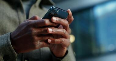 A man of African American appearance uses a mobile phone, standing near a building in which lamps shine in the windows. Close-up of male hands with a phone, no face. in 4k, red komodo video