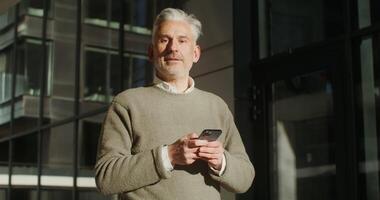 A gray-haired man dressed in a warm sweater is distracted from the mobile phone in his hands and looks at the camera with a smile, standing near the city building. in 4k, red komodo video